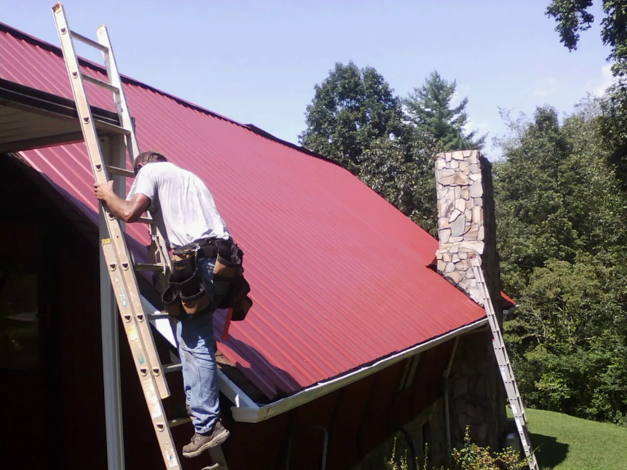 Roofer on a ladder beside a red metal roof and stone chimney — Soffit & Fascia Repair by Roofing Brothers in Wallace